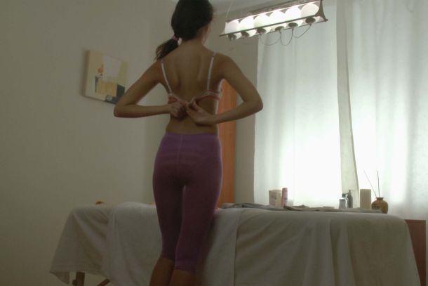 Model Milana F leaning over a bed topless and happy.